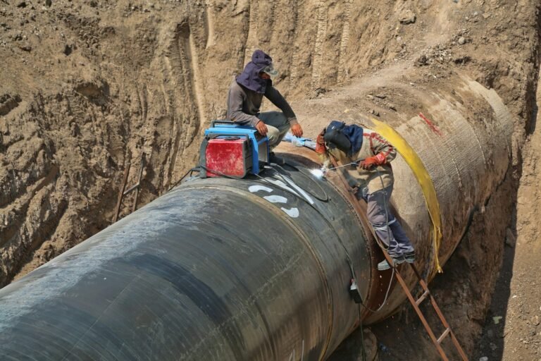 Workers inside a deep excavation trench beside a large pipeline